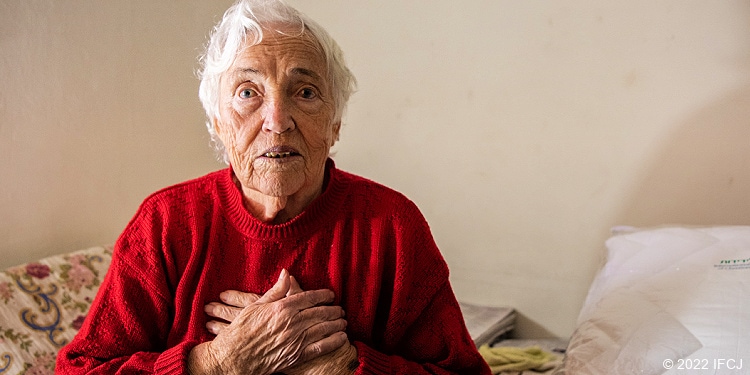 Elderly Jewish woman holding hands to chest in red sweater.