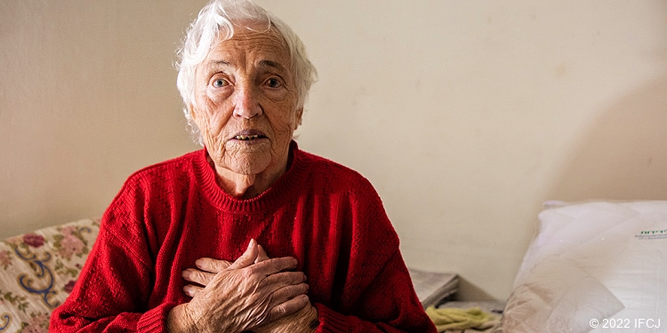 Elderly Jewish woman holding hands to chest in red sweater.