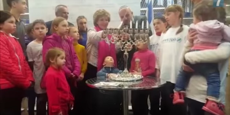 A woman lighting a menorah with sixteen other people as some of them are wearing IFCJ branded shirts.