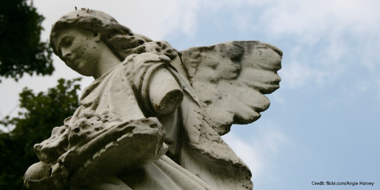 Close up image of the top half of an angelic statue against trees and a cloudy blue sky.