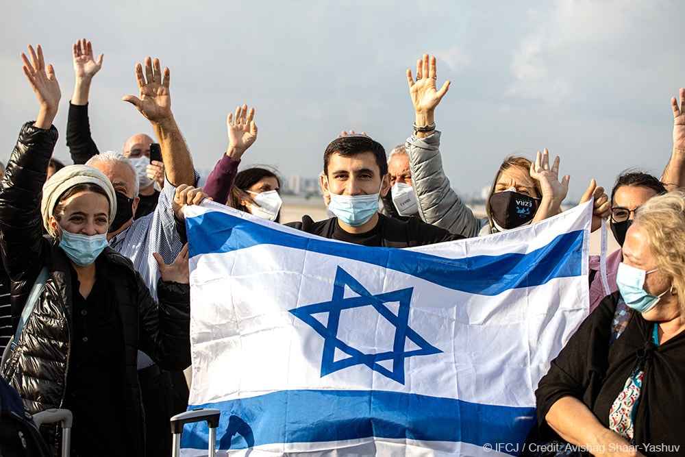 Group of olim standing on tarmac with hands raised and holding flag of Israel
