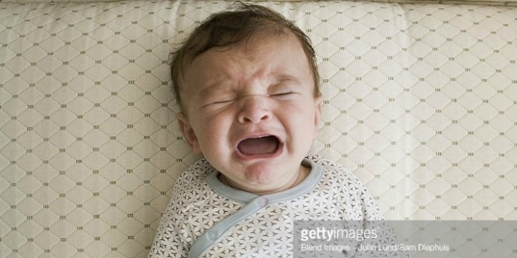 A baby in a white onesie crying in a white crib.