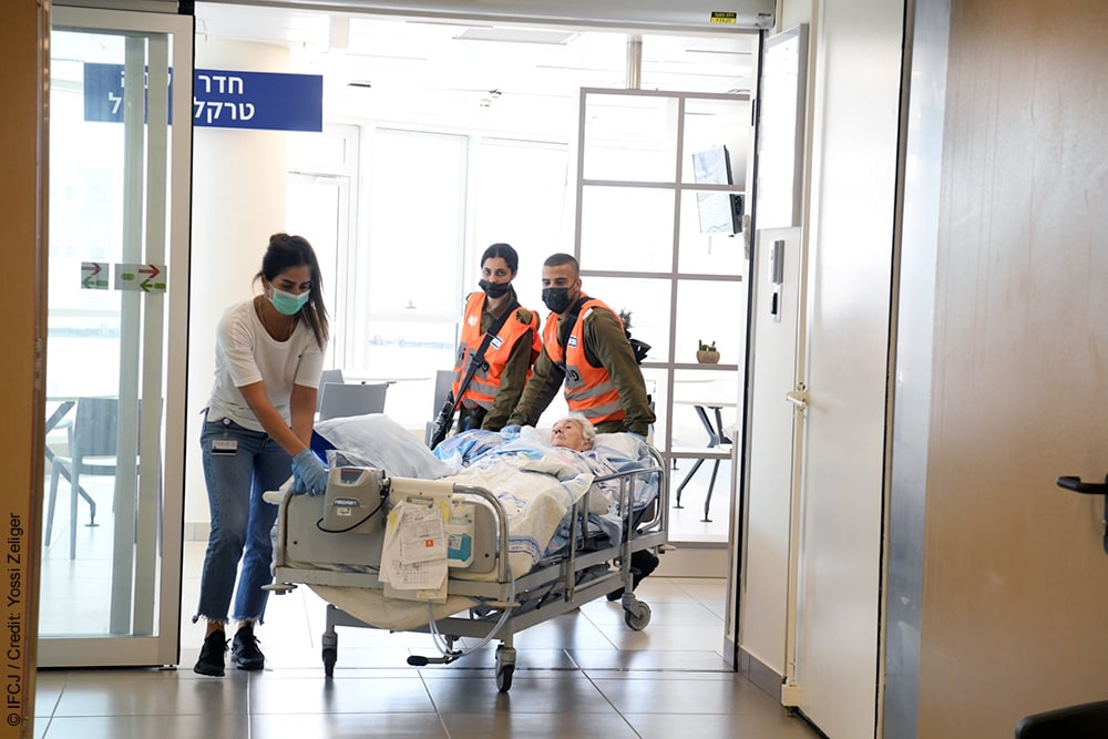 3 people pushing a medical cart with an elderly Jewish person on it.