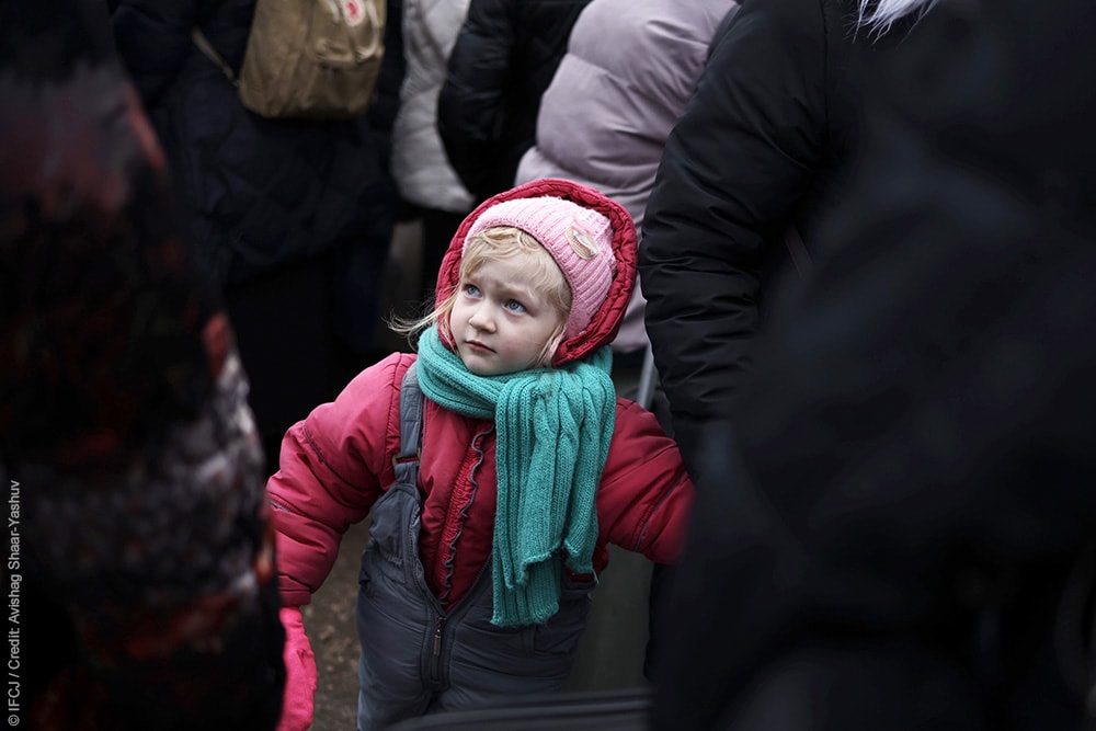 Little girl wearing winter clothing stands in the middle of a group of people holding someones hand.
