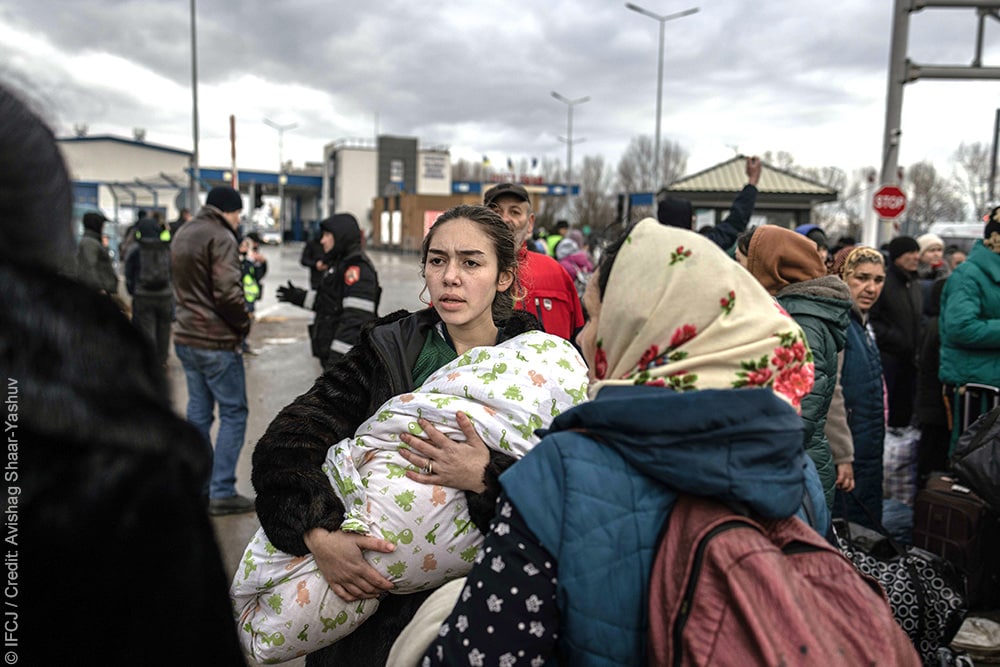 Adult woman holding infant wrapped in a blanket, surrounded by other refugees, walking through the street.