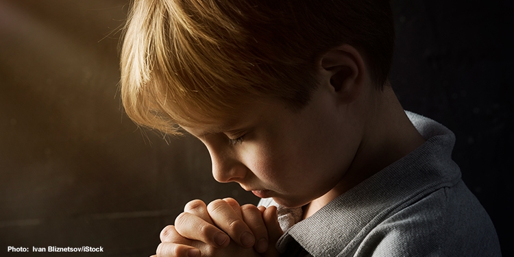 Young boy in a gray shirt bowing his head down while his hands are folded in prayer.