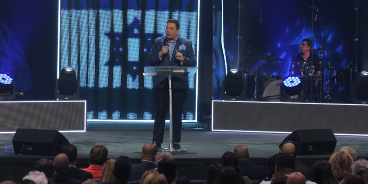 Carols Ortiz standing at a glass podium giving a sermon on stage.