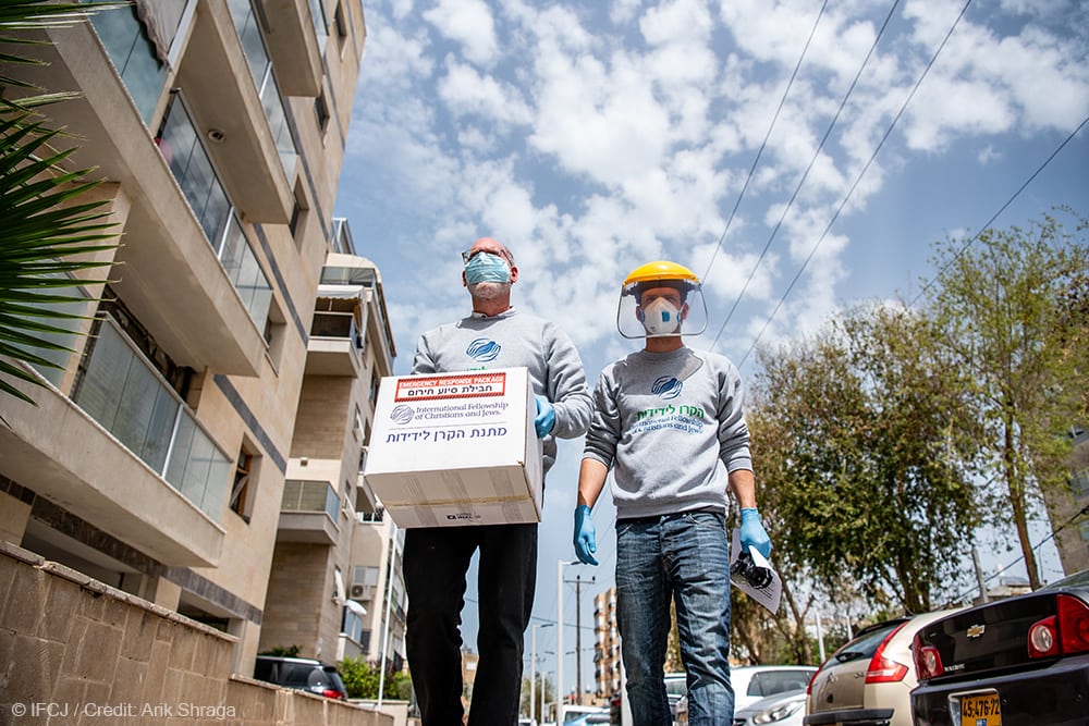 Two IFCJ staff members in masks and gloves stand in the street carrying a food box