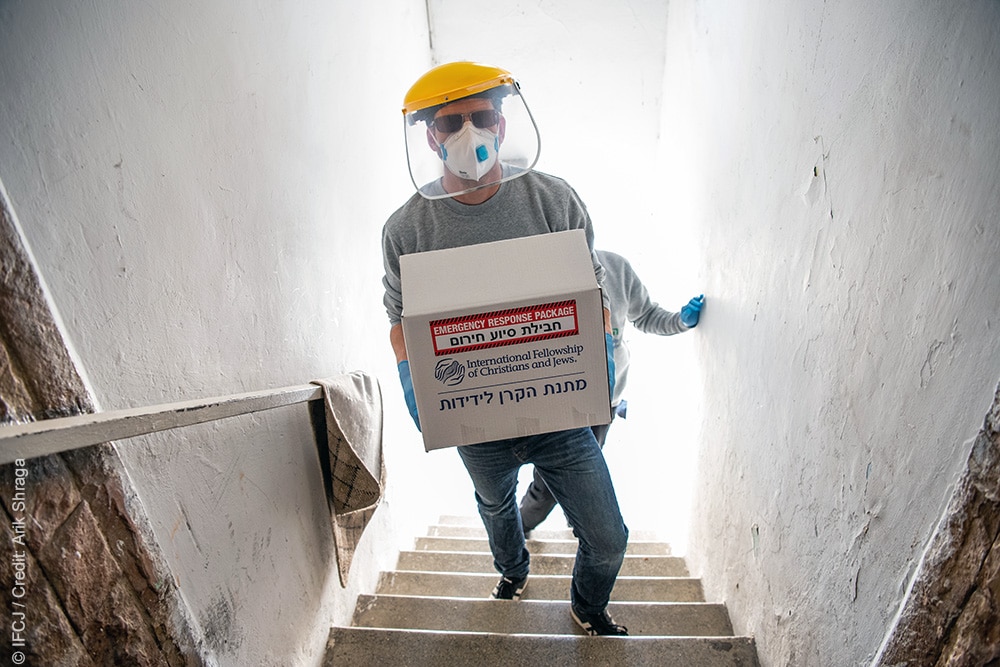 IFCJ staff person in mask and gloves climb stairs carrying food box.