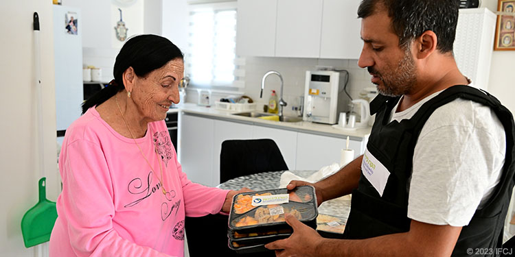 adult man in IFCJ-branded flak jacket delivering food to elderly woman