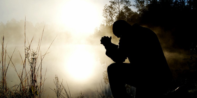 Person bowing their head with their hands clasped praying next to a lake