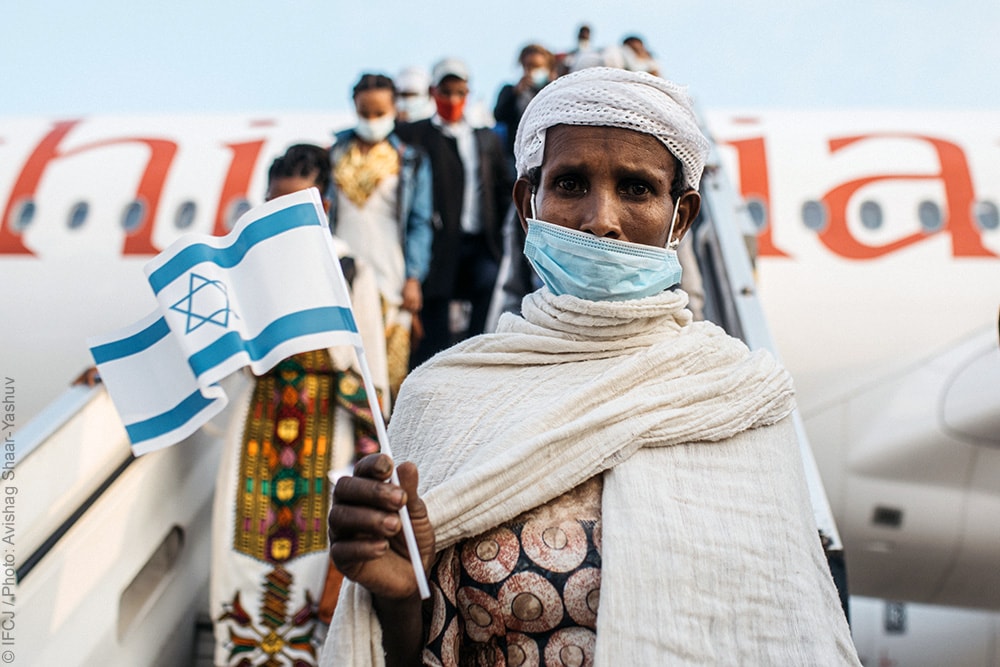 Elderly woman wearing white headscarf and shawl waves flag of Israel standing in front of an airplane