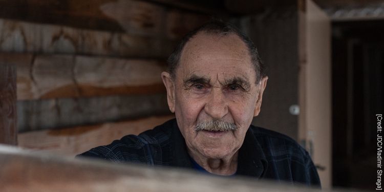 Semyon, an elderly male IFCJ recipient standing in a room covered with wooden walls.