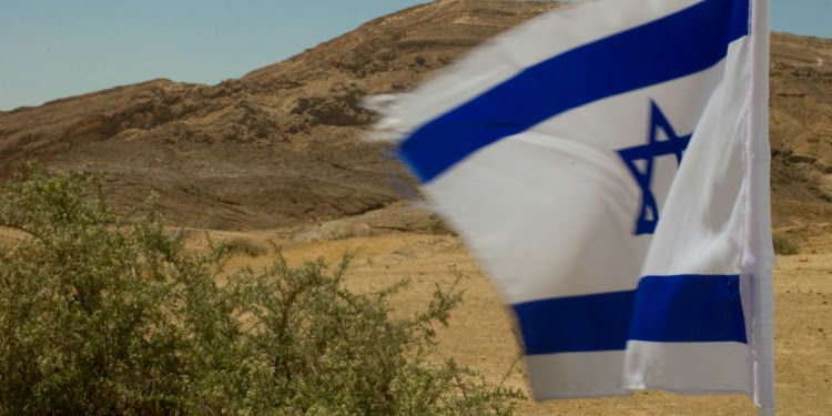 Israeli flag flying in the air with a desert scenery behind it.