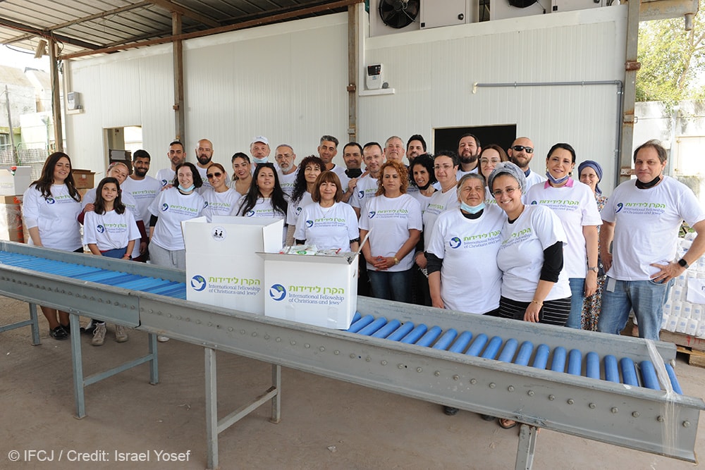 Group of people stand behind a conveyer belt packing food boxes at Mifal HaHesed soup kitchen