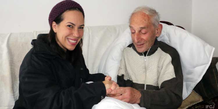 Yael Eckstein holding hands with an elderly Jewish man sitting on a couch.