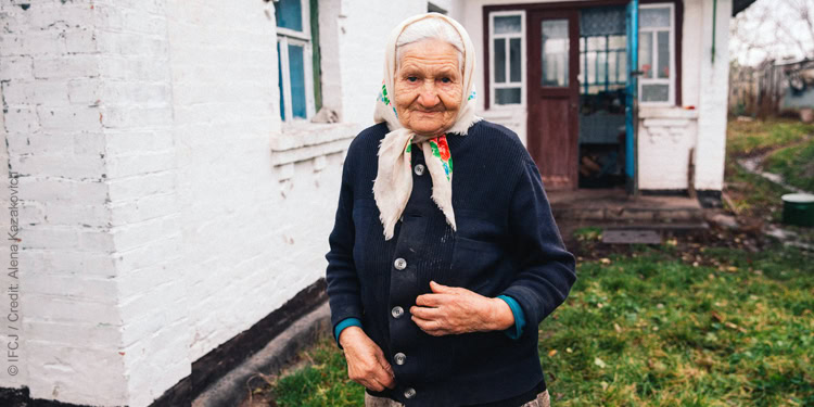 Elderly woman, standing outside a white house, wearing a blue sweater.