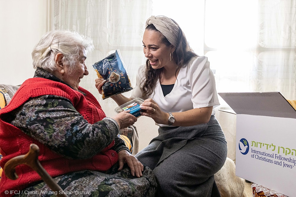 Elderly woman sits with Yael Eckstein holding packaged food.