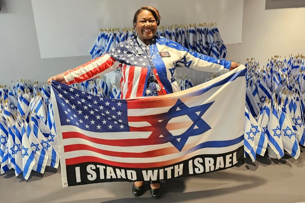 Woman stands holding a flag that is half the US flag and Israel flag. At the bottom of the flag are the words "I Stand With Israel"