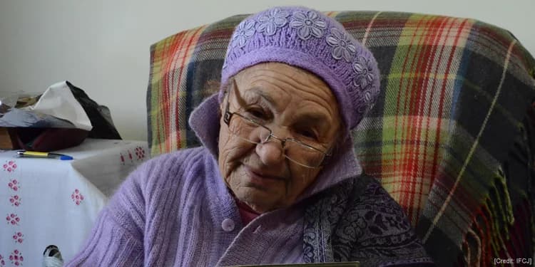 Elderly Jewish woman in a matching purple beanie and sweater sitting on a patterned chair.