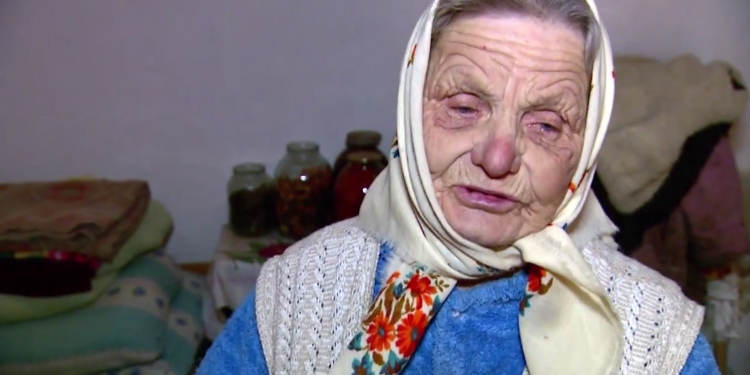 Elderly Jewish woman in a white floral-patterned headscarf looking down while several blankets and jars are behind her.