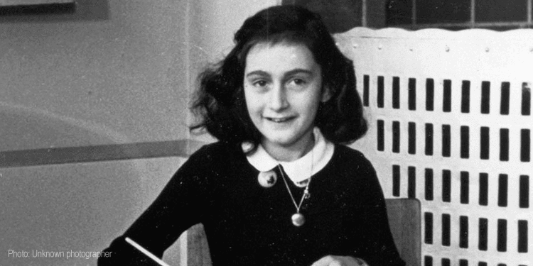Image of Anne Frank sitting at a table holding a pencil