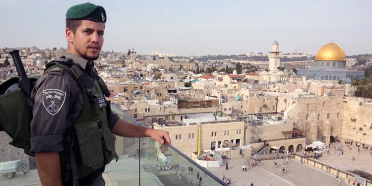 A soldier in full uniform with his hand on a rail overlooking the city of Jerusalem.