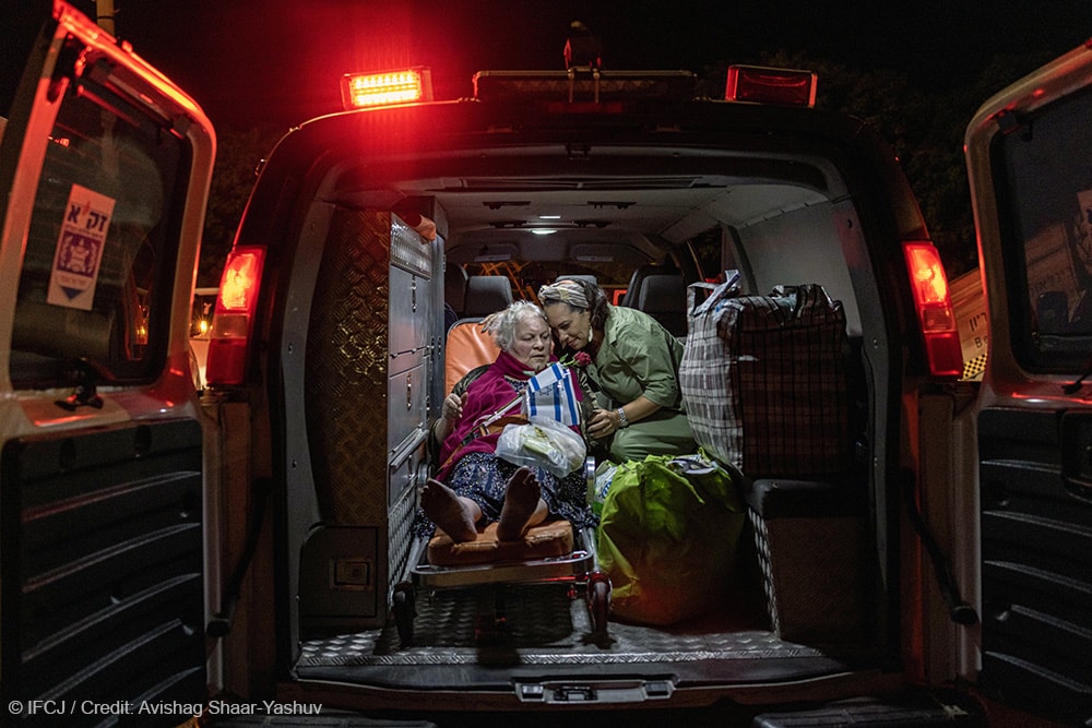 Yael Eckstein sits in the back of ambulance hugging an elderly woman on a gurney with her belongings in bags around her