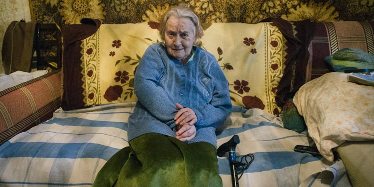 Elderly Jewish woman in a blue sweater sitting on a bed with her arms crossed.