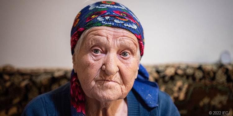 Elderly woman wearing blue scarf sit on couch looking at camera