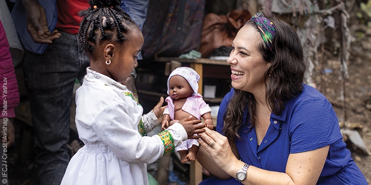 Yael kneels down to hand a doll to a young girl.