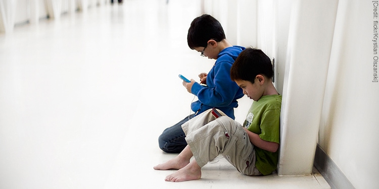 Two young boys sitting on a white floor while playing games on their cellphones.