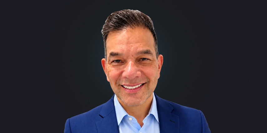 Retrato del Dr. Carlos Ortiz, sonriendo con un traje azul y una camisa azul claro sobre un fondo oscuro.
