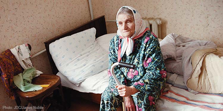 Elderly woman sitting on bed holding a cane, green patterned dress robe, white head scarf wrap
