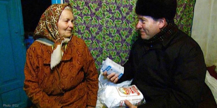 Rabbi Eckstein in winter clothing sitting next to an elderly Jewish woman handing her food.