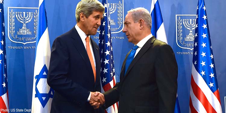 Two men shaking hands while in front of Israeli and American flags.