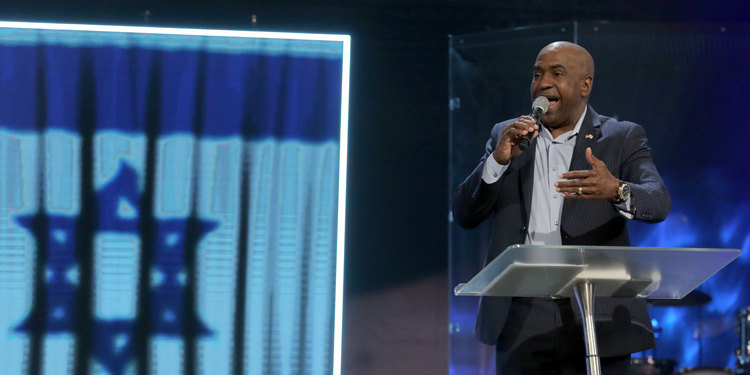 Roger Cheeks giving a sermon on a stage at a glass podium with the Israeli flag behind him.