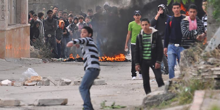 A young boy with his hand cocked back with crowds of another people behind him amongst a dirty road and fires.