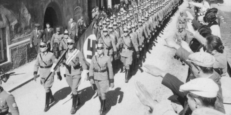 Black and white image of Nazi soldiers walking alongside a flag with the swastika.