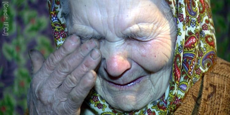 Elderly Jewish woman in a paisley head scarf looking down wiping her tears.