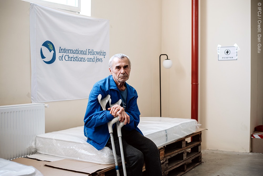 Elderly man sits on a matress supported by wooden pallets while holding to crutches