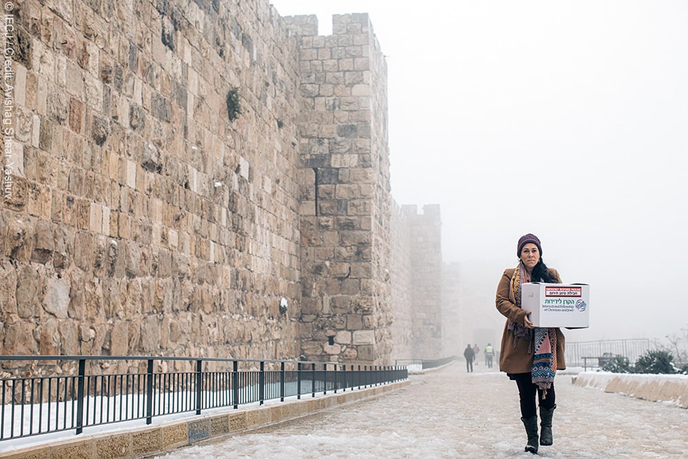 Snow in Jerusalem, Yael Eckstein holding a branded logo emergency response package, walking towards camera, old wall to the side of her, snow on the ground