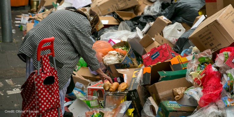 An elderly woman grabbing bread from a pile of trash.