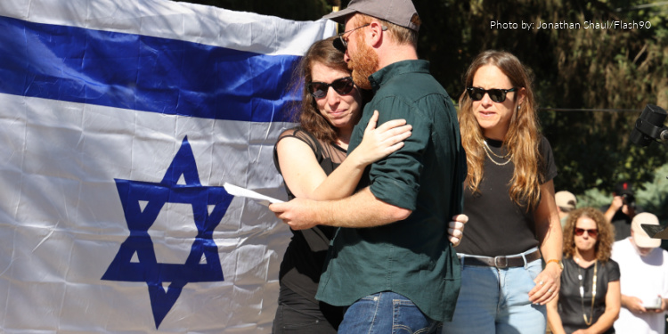 Woman crying in the arms of a man with the Israeli flag behind her.