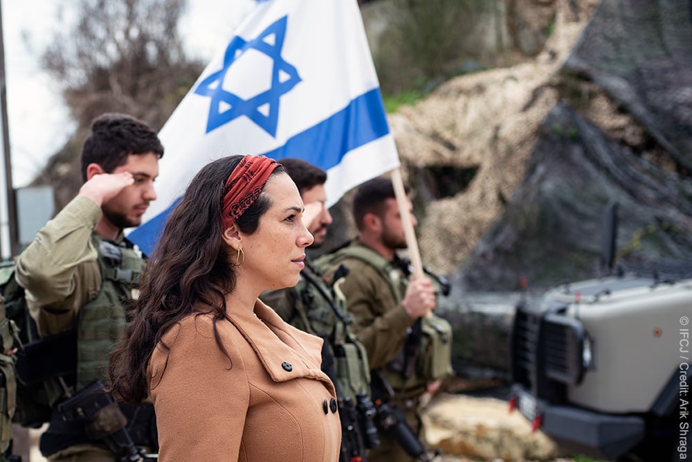 Yael looking forward while standing next to soldiers holding the Israeli flag.
