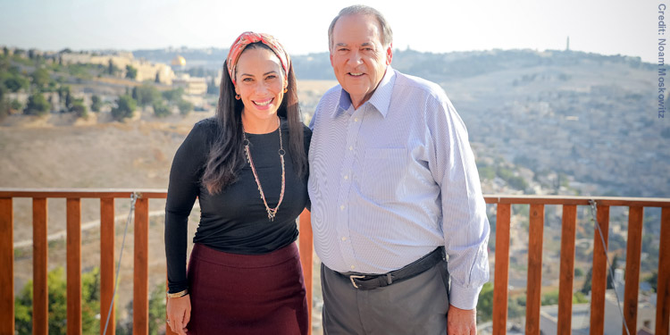 Yael Eckstein and Mike Huckabee stand next to a fence with Israel behind them in the landscape.