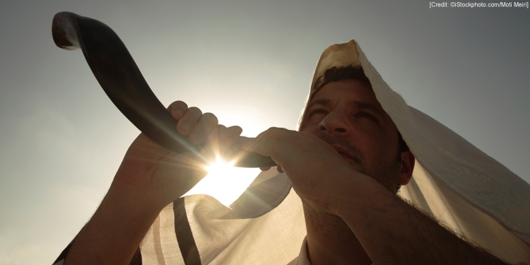 A man in a white head covering blowing a shofar on a sunny day.