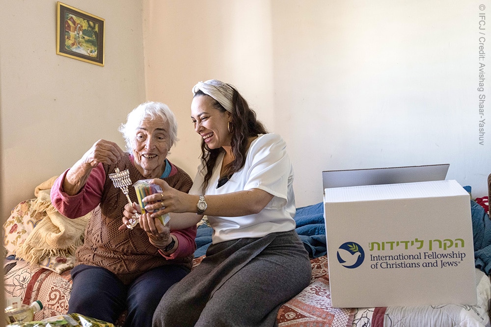 Zoya, elderly woman, sits on a bed with Yael Eckstein holding menorah and candles next to a white box with the IFCJ logo printed on it.